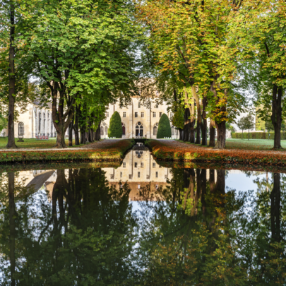 Un chemin bordé d'arbres avec le Bâtiment des moines Abbaye de Royaumont se reflétant dans un étang, entouré de feuillages d'automne.